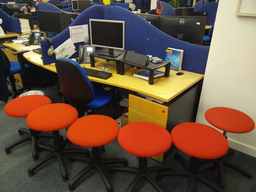 Photo of desk in typical office from the 2010s, with a desk phone, monitor, cradle for laptop, surrounded by red stools for no obvious reason